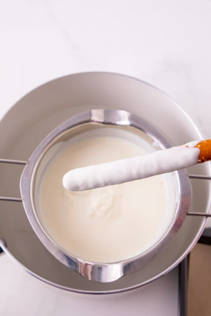 Stainless mixing bowl filled with white melted chocolate and a chocolate covered pretzel resting on the rim