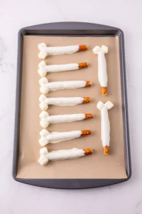 Baking tray lined with parchment holding white chocolate-coated pretzel bone sticks arranged in rows.