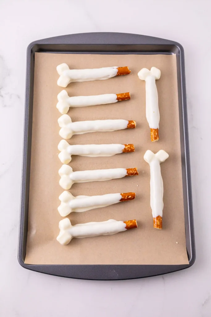 Baking tray lined with parchment holding white chocolate-coated pretzel bone sticks arranged in rows.