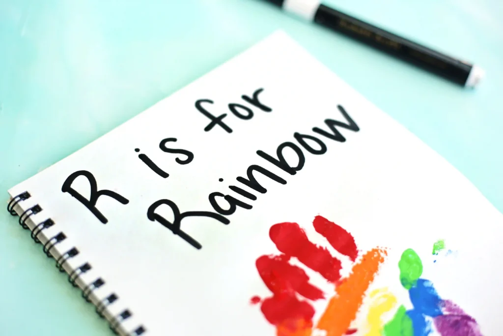 Spiral notebook page with the handwritten phrase'R is for Rainbow' and rainbow paint splashes on a light turquoise background.