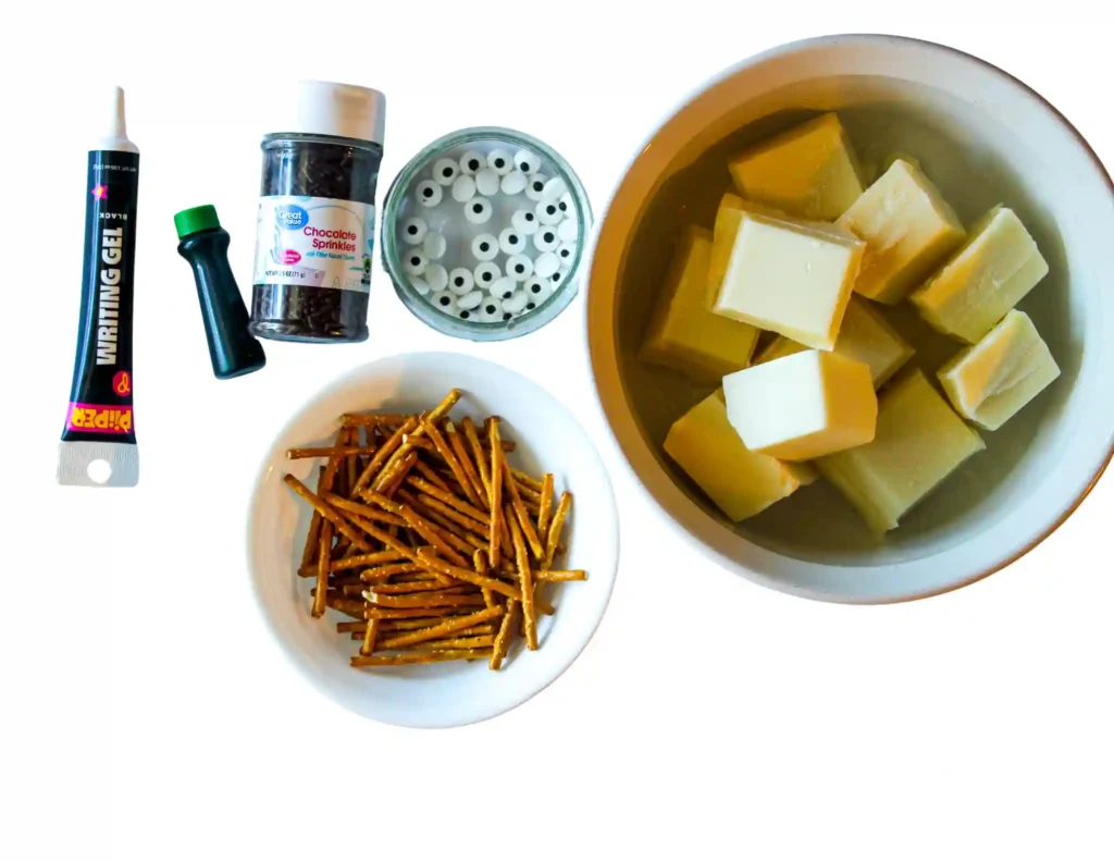 ingredients for the frankenstein bark: white chocolare chunks in a bowl, pretzel sticks, a jar of chocolate sprinkles, a tub of edible pearls, and an icing tube on a white background.