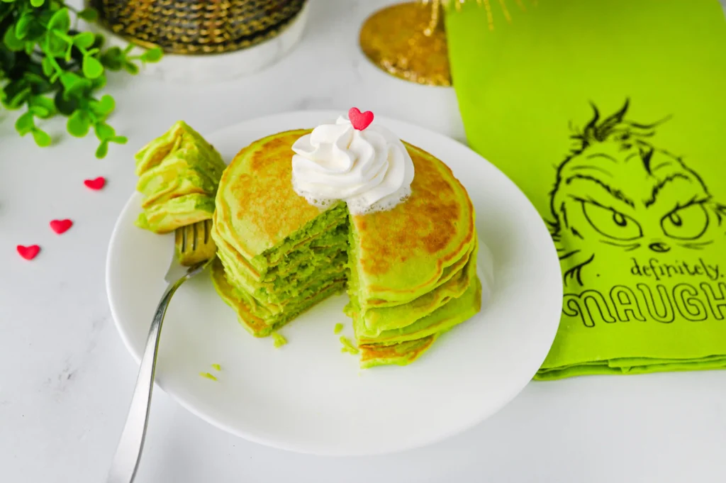 green panckaes with whipped cream and heart sprinkles on a white plate next to a grinch napkin
