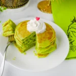 green panckaes with whipped cream and heart sprinkles on a white plate next to a grinch napkin