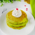 green panckaes with whipped cream and heart sprinkles on a white plate next to a grinch napkin