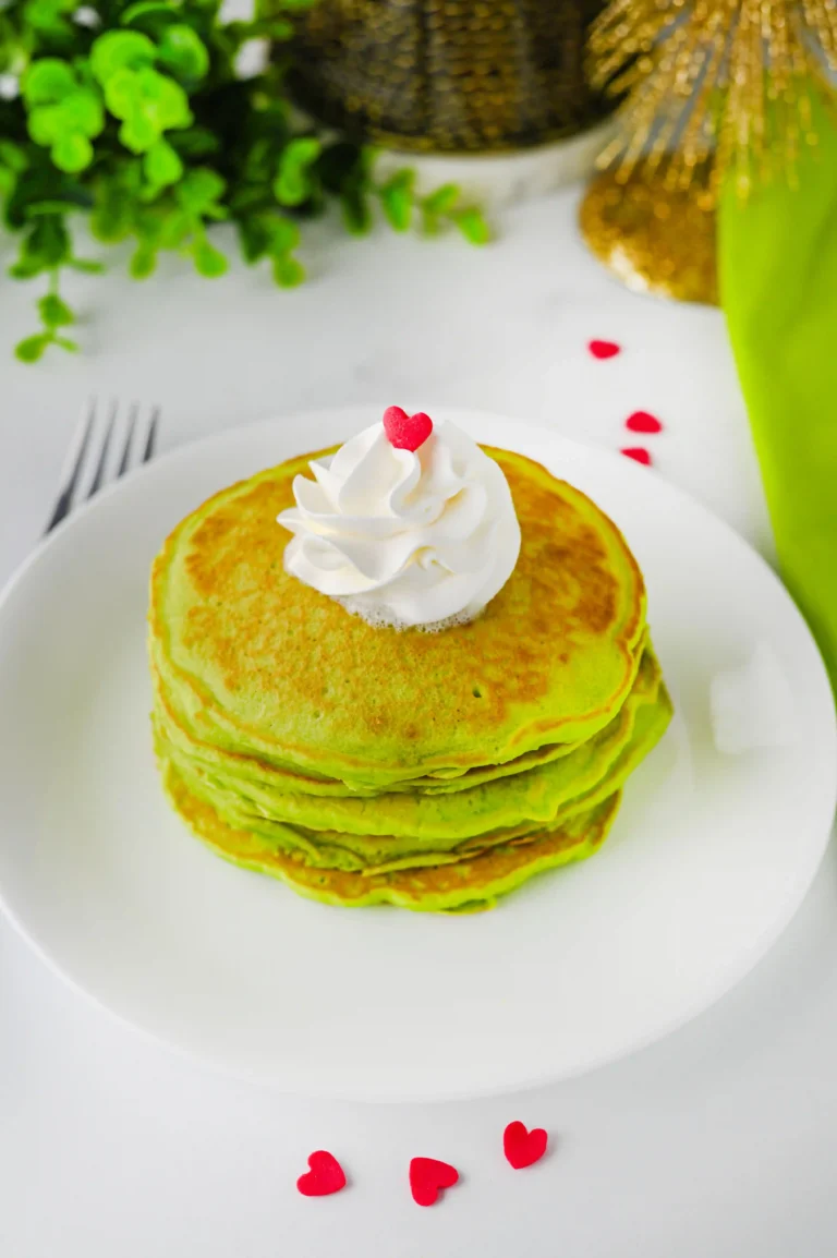 green panckaes with whipped cream and heart sprinkles on a white plate next to a grinch napkin