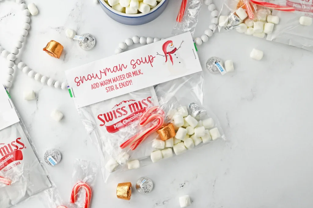 Packet of Swiss Miss milk chocolate cocoa mix with marshmallows and wrapped candies scattered on a white surface, suggesting hot chocolate prep.