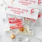 Assorted holiday treats around Swiss Miss hot chocolate packets on a white surface, including candy canes and foil-wrapped candies.