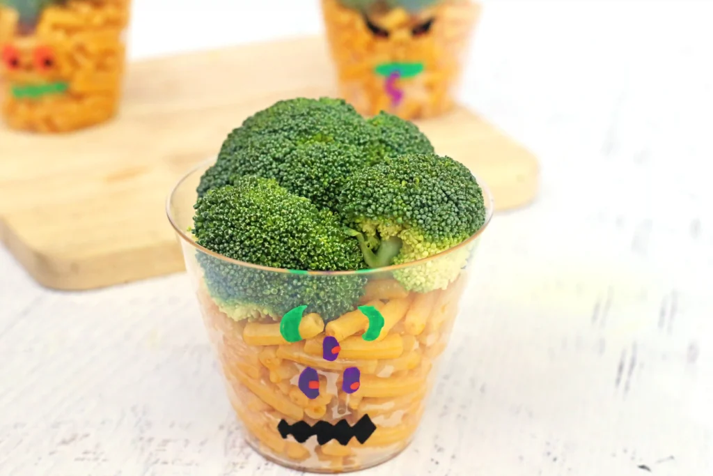 a clear cup with a monster face drawn on with mac and cheese and broccoli inside