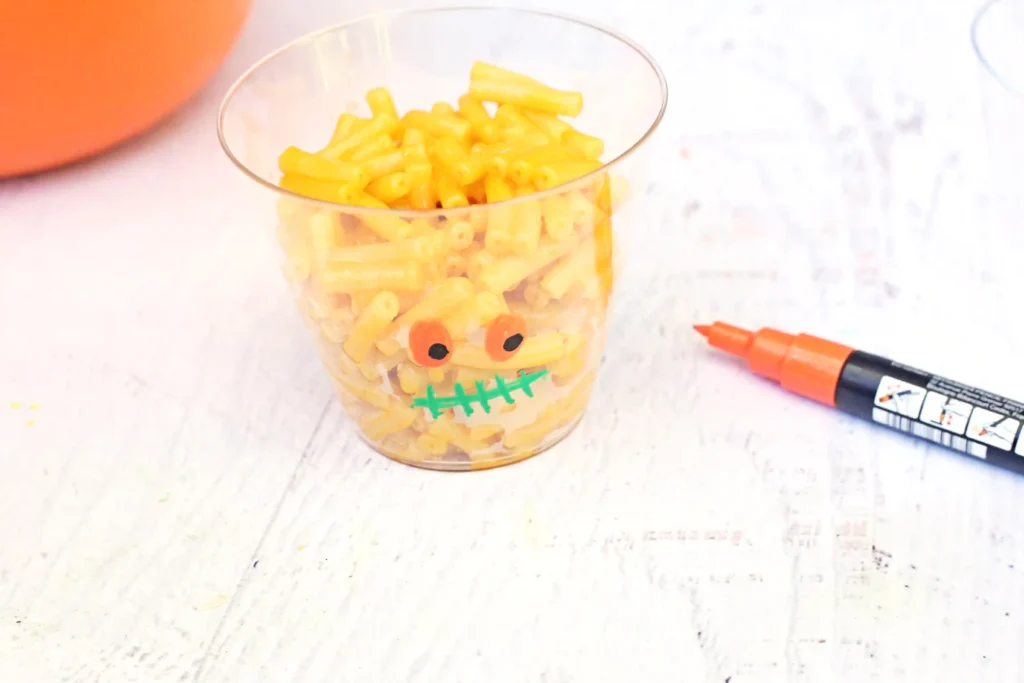 A clear cup filled with yellow macaroni shaped pasta, decorated with drawn eyes and a mouth to resemble a face, with an orange marker nearby on a light surface.