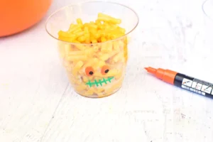 A clear cup filled with yellow macaroni shaped pasta, decorated with drawn eyes and a mouth to resemble a face, with an orange marker nearby on a light surface.