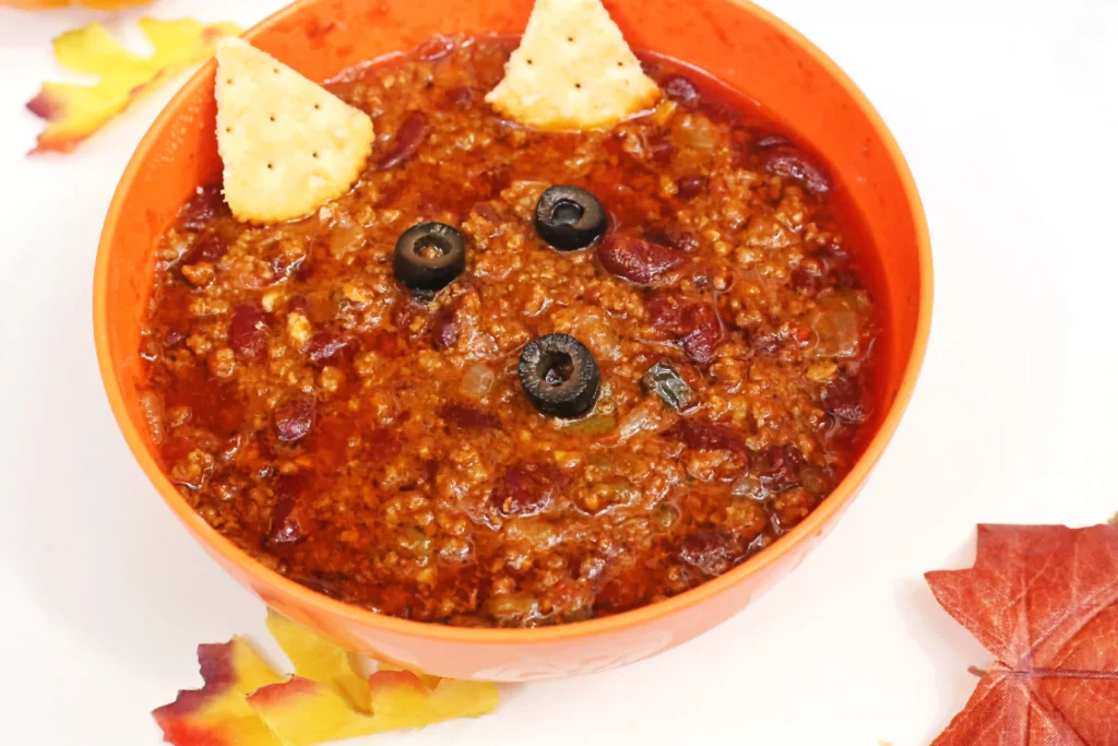 Orange bowl of chili with black olive slices and tortilla chips on top, set on a light surface with autumn leaves nearby.