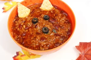 Orange bowl of chili with black olive slices and tortilla chips on top, set on a light surface with autumn leaves nearby.