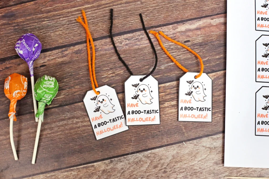 Halloween gift tags with a cute ghost and the text ‘Have a Boo-tastic Halloween!’ on a wooden surface beside colorful lollipops.