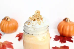 Milkshake topped with whipped cream and crumbled cookies in a glass, flanked by mini pumpkins on an autumn-themed table.
