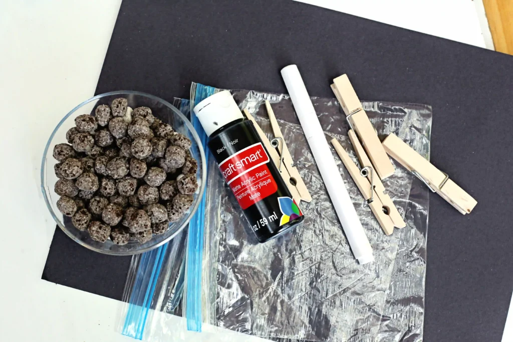 Bat Snack Bags - Cultivating Brilliant Minds supplies for the bat snack bags: a bowl of small brown round beads or cereal-like pieces beside a black spray paint can and several wooden clothespins on a metallic, textured surface.