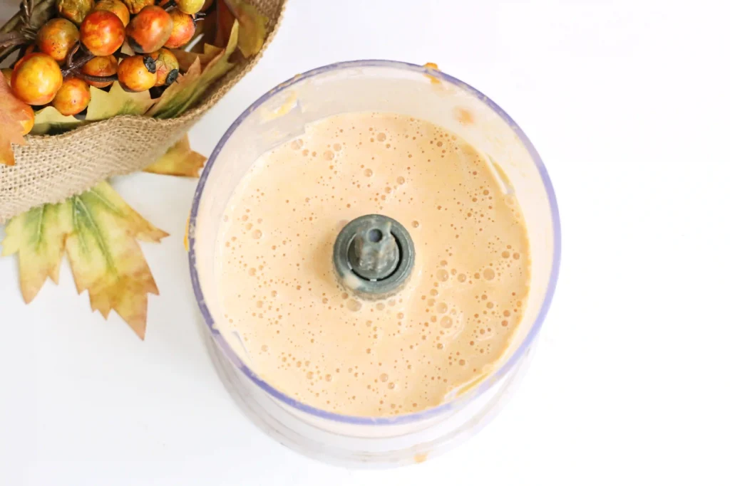 Blender with creamy pumpkin smoothie on a white surface, surrounded by a basket of small orange fruits and autumn leaves.