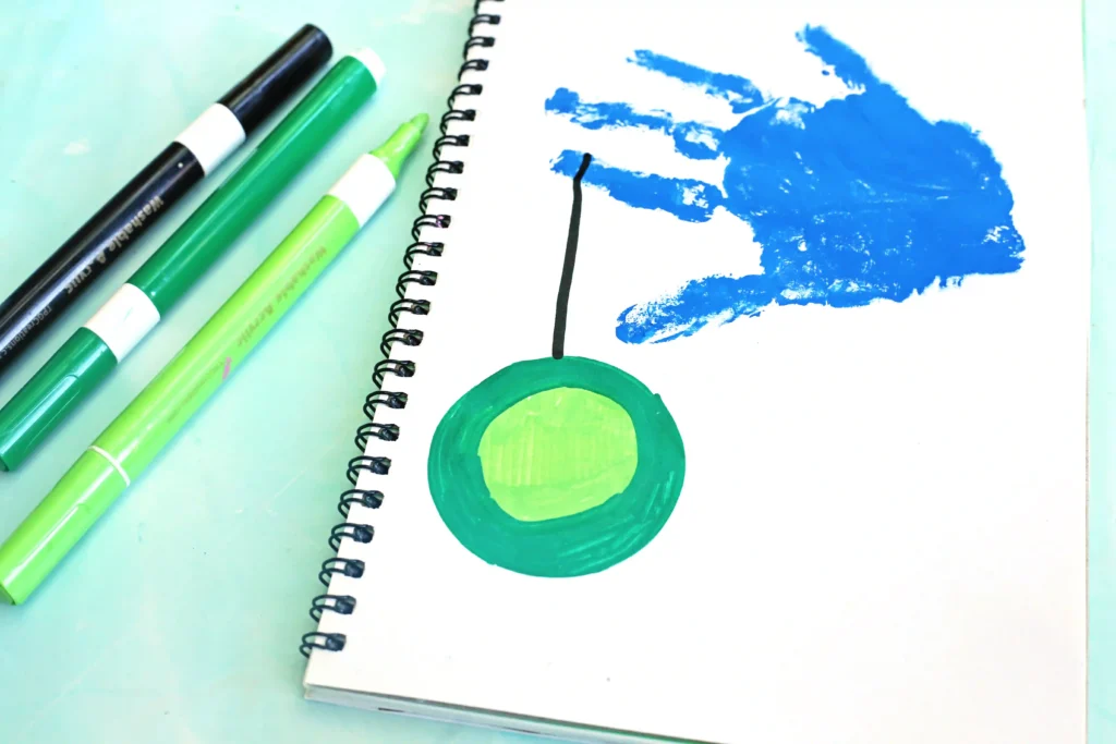 Spiral notebook page with a bold blue handprint and a yo yo, beside green markers on a pale surface.