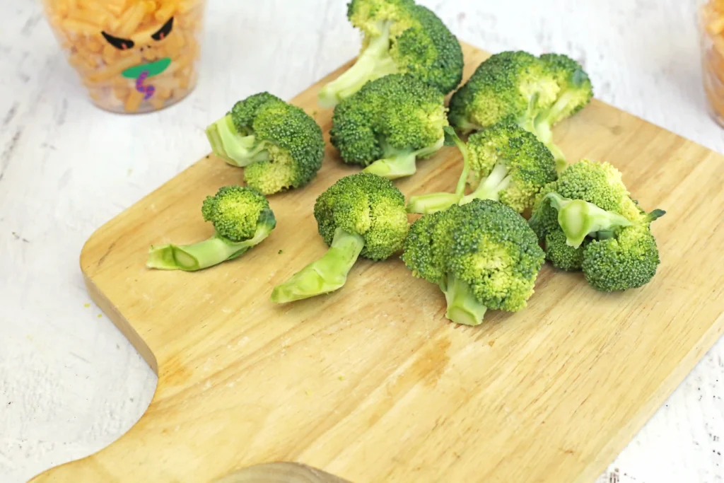 Broccoli florets scattered on a wooden cutting board with a blurred container of fries in the background, ready for cooking.