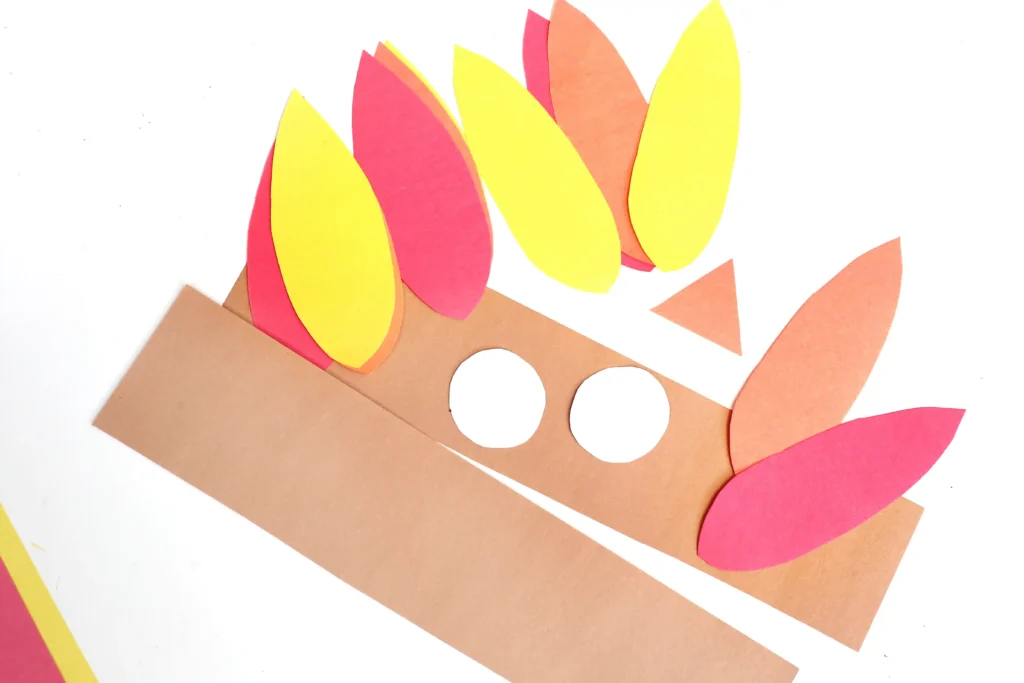 Brown construction paper base with white circular cutouts, layered yellow, pink, and orange teardrop shapes arranged like feathers.