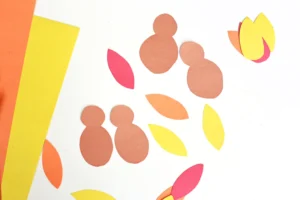 Abstract paper collage with orange, pink, and yellow shapes on a white background resembling leaves and rounded figures.