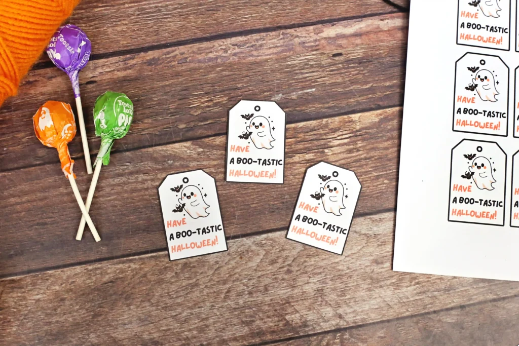 Halloween-themed tags with a cute ghost and bats reading'Have a Boo-tastic Halloween!' scattered on a wooden surface, beside wrapped lollipops and an orange sleeve partially visible on the left.