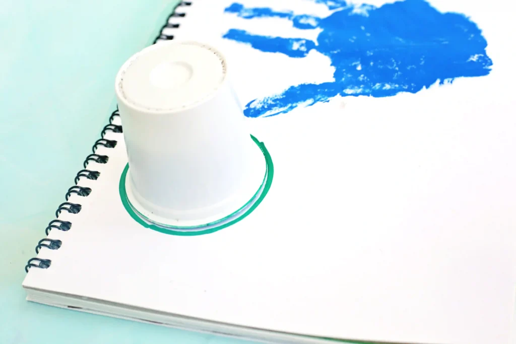 Spiral-bound notebook opened on a light blue surface with a white coffee pod on top and a blue paint smear on the page.