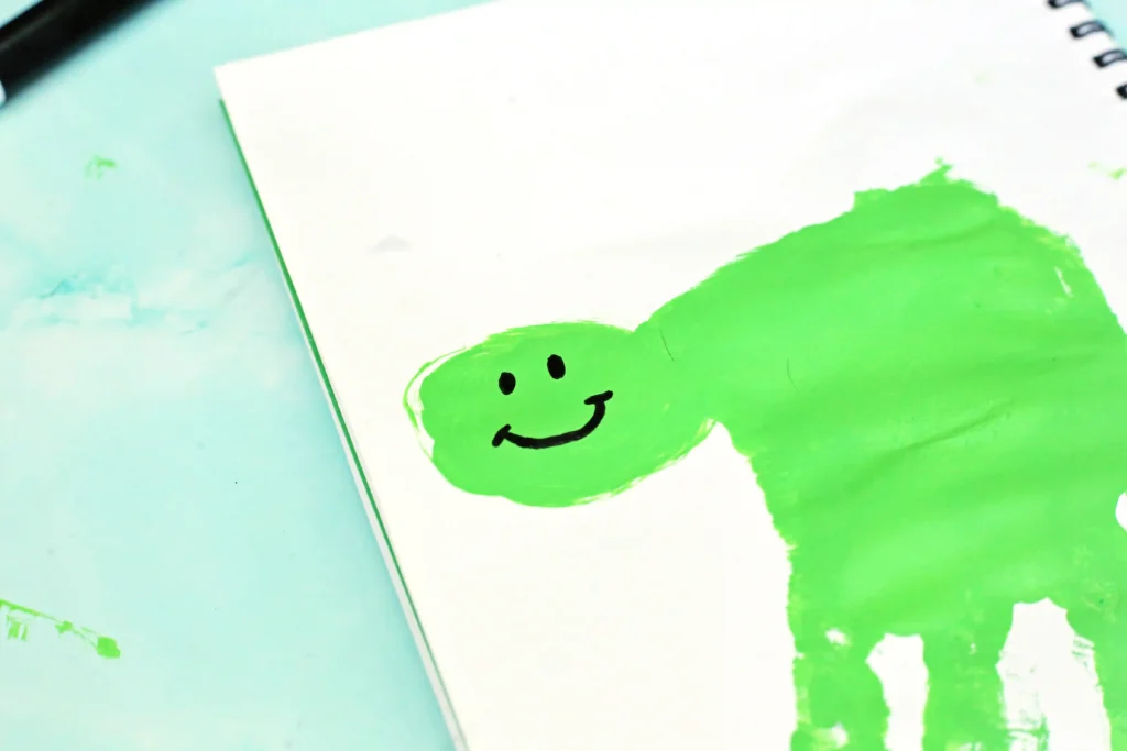 Close-up of lime-green paint handprint on white paper with a small black smiley face, and a notebook edge visible on the right side.