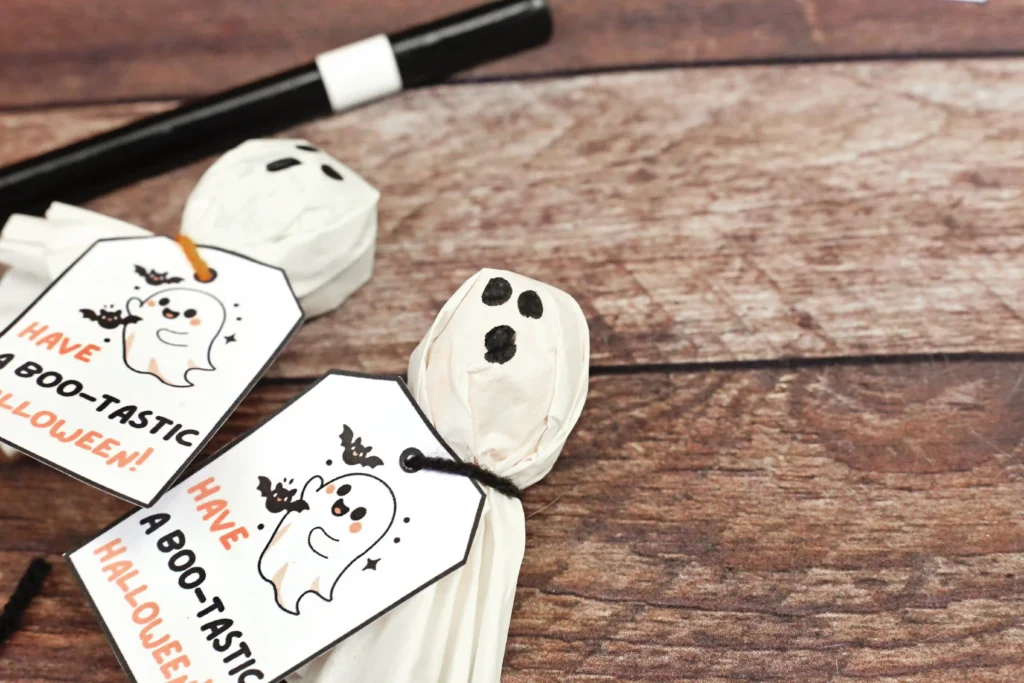 Close-up of paper ghost face on a wooden tabletop with Halloween tags reading'Have a Boo-tastic Halloween'.