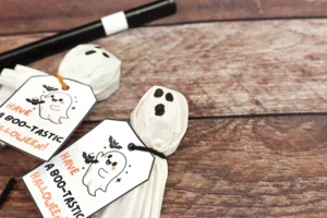 Close-up of paper ghost face on a wooden tabletop with Halloween tags reading 'Have a Boo-tastic Halloween'.