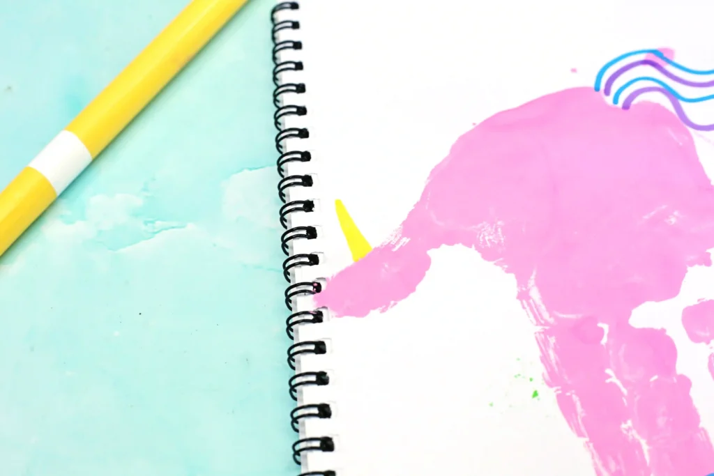 Spiral notebook with a pink handprint and a yellow pencil on pastel blue and pink watercolor background.