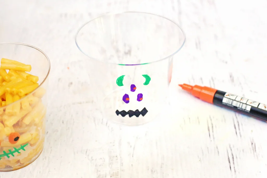 A clear glass cup decorated with a colorful face drawn in green, purple, and black markers, with a bowl of macaroni shapes to the left and an orange marker to the right.
