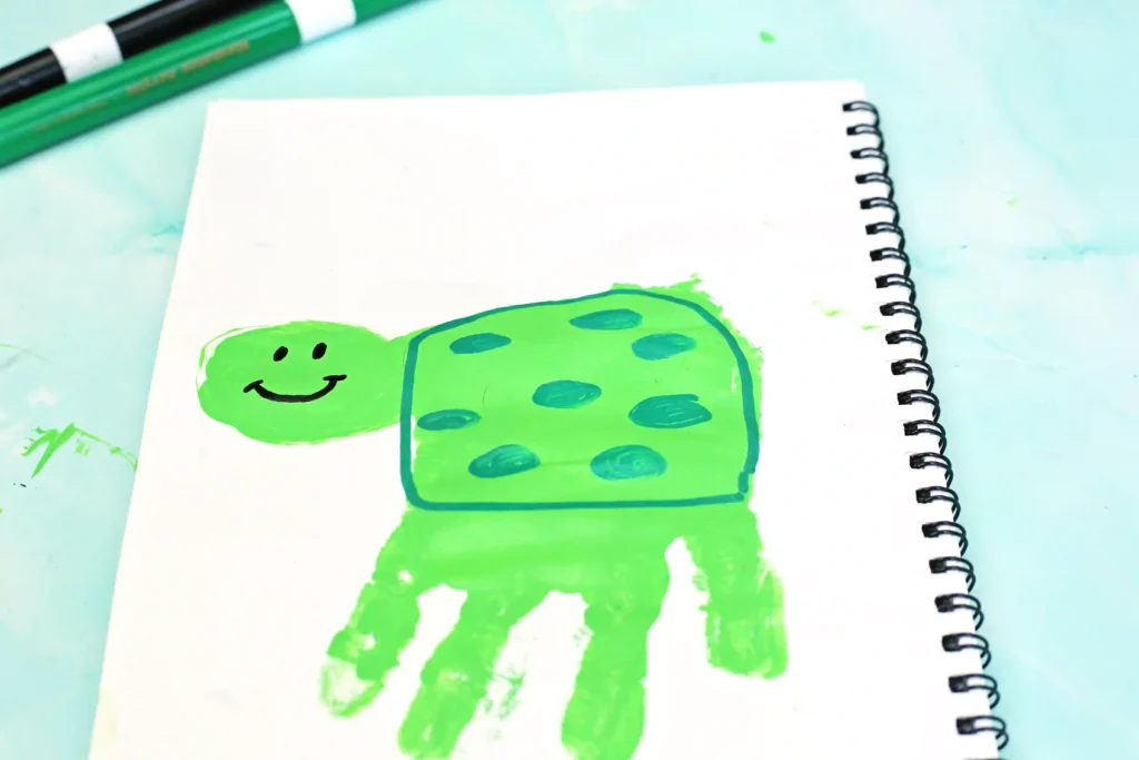 Green cartoon turtle drawn with markers on a white spiral notebook; two green markers lie at the top.