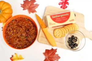 Fall-themed snack spread: chili in an orange bowl, cheddar cheese, crackers, and autumn leaves on a white surface with a wooden board nearby.