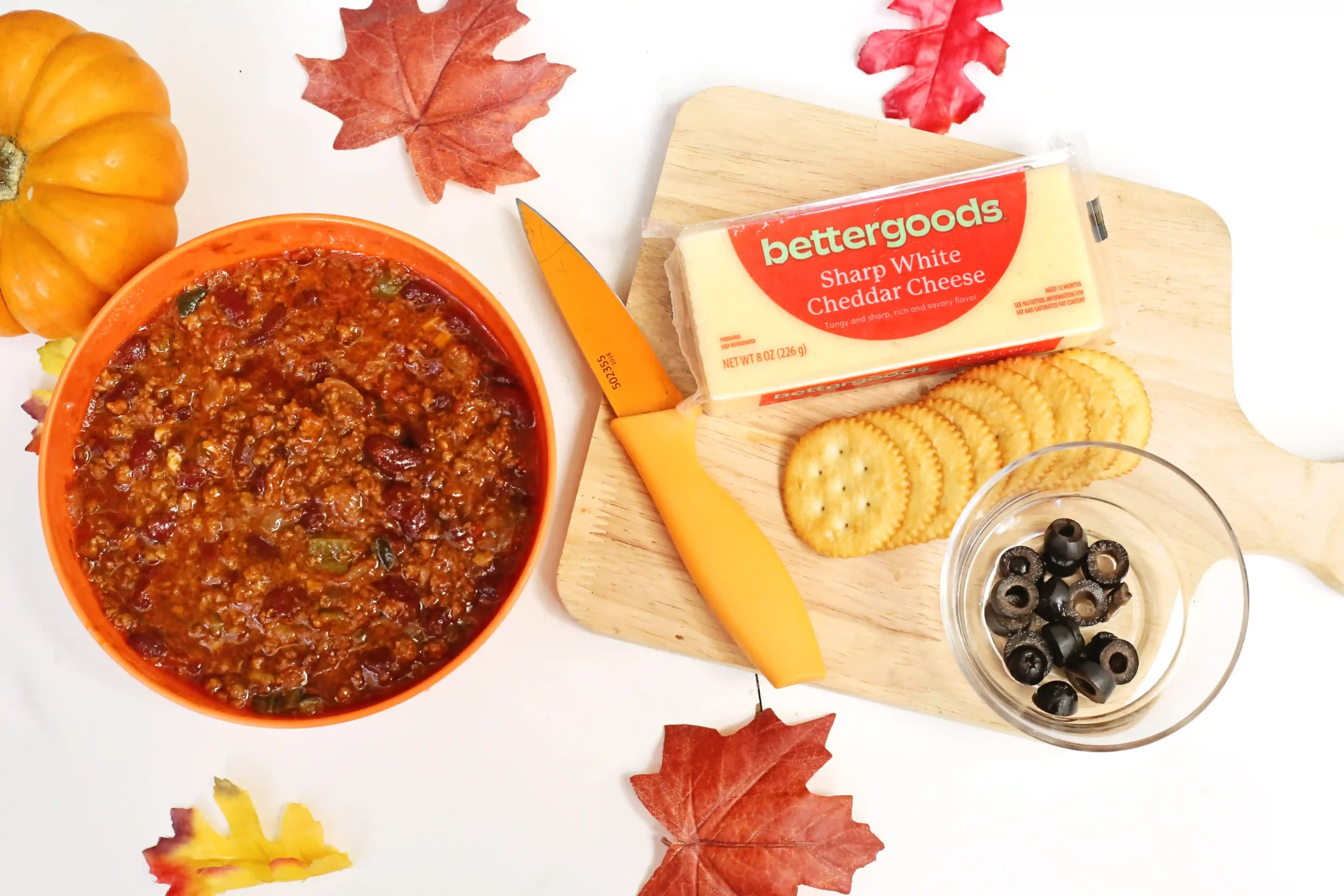 Fall-themed snack spread: chili in an orange bowl, cheddar cheese, crackers, and autumn leaves on a white surface with a wooden board nearby.