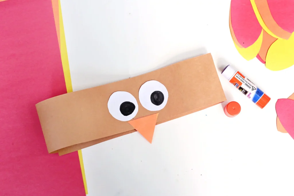 Brown paper craft with two large white eyes and an orange triangular beak on a white surface, with a glue stick nearby and colorful cutouts around.