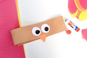 Brown paper craft with two large white eyes and an orange triangular beak on a white surface, with a glue stick nearby and colorful cutouts around.