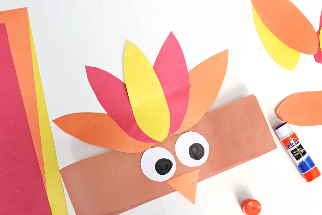 Colorful paper collage of a cartoon turkey with orange head, yellow and pink feathers, and large googly eyes on a white background.