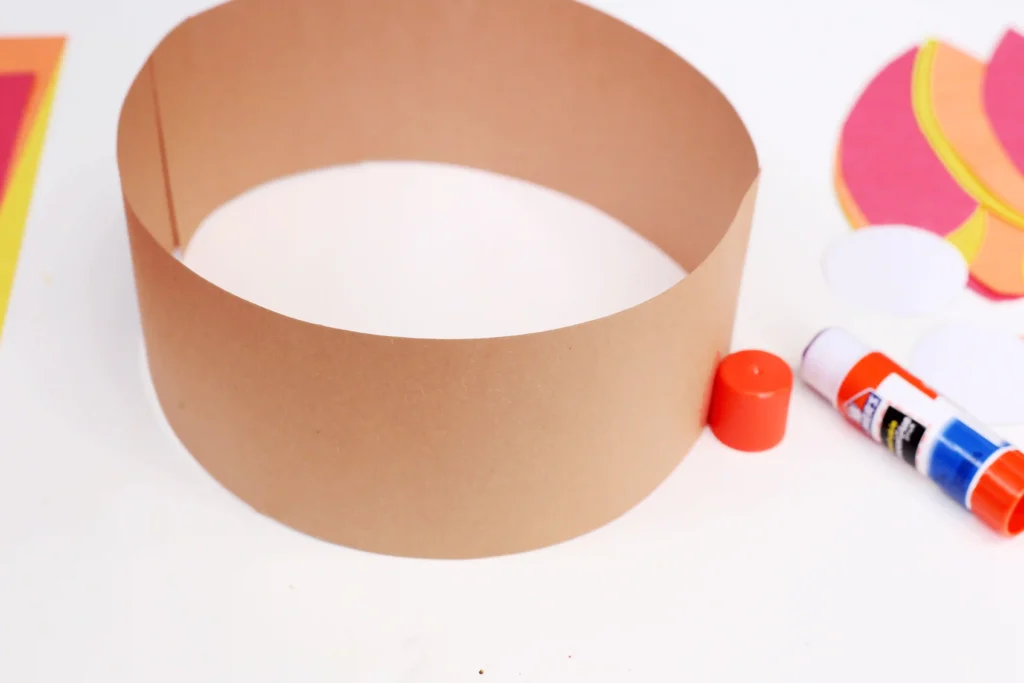 Close-up of a paper craft featuring a brown hollow ring surrounded by colorful paper shapes and glue sticks on a white surface