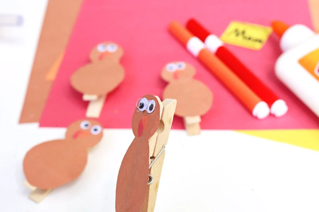 Close-up of a brown clothespin turkey with googly eyes among other turkey crafts on a pink background menagerie of holiday crafts.