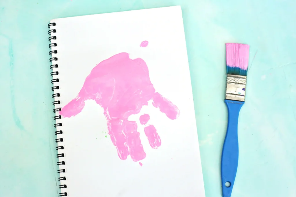 White spiral notebook with pink paint handprint on its page, lying on a turquoise surface with a blue and pink paintbrush nearby.