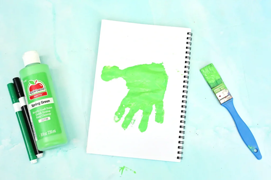 Green craft supplies scene: a bottle of lime paint, two markers, and a spiral notebook with a bright green handprint on its page against a turquoise surface.