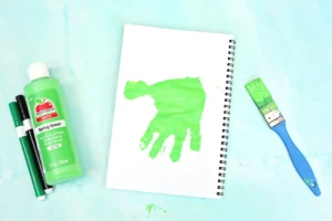 Green craft supplies scene: a bottle of lime paint, two markers, and a spiral notebook with a bright green handprint on its page against a turquoise surface.