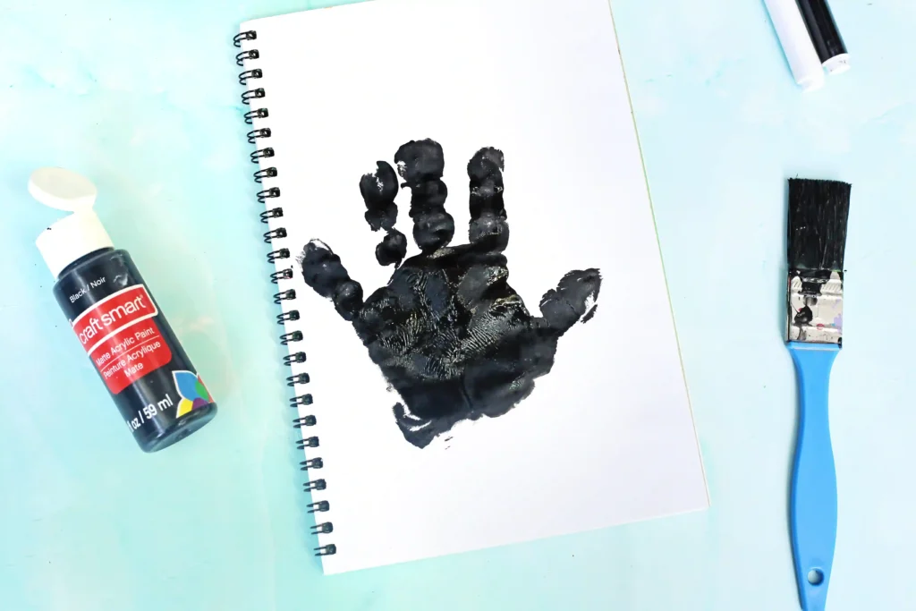 A spiral-bound notebook opened to a page with a black paint handprint, surrounded by art supplies on a light blue surface: a bottle of black acrylic paint, a paintbrush, and a blue-handled tool nearby.