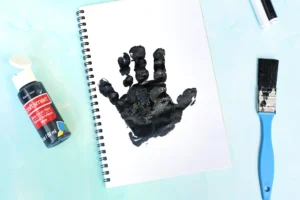 A spiral-bound notebook opened to a page with a black paint handprint, surrounded by art supplies on a light blue surface: a bottle of black acrylic paint, a paintbrush, and a blue-handled tool nearby.