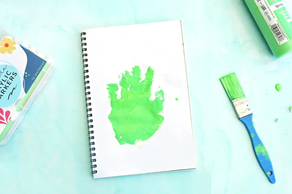 Spiral notebook with a green paint smear on a white page, surrounded by art supplies on a turquoise surface.