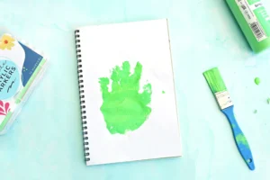 Spiral notebook with a green paint smear on a white page, surrounded by art supplies on a turquoise surface.