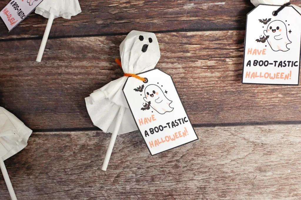 Ghost-themed Halloween printable tags attached to a ghost lollipop on a wooden background with the text Have a Boo-tastic Halloween! on white cards.