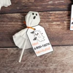 Ghost-themed Halloween printable tags attached to a ghost lollipop on a wooden background with the text Have a Boo-tastic Halloween! on white cards.