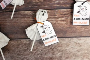 Ghost-themed Halloween printable tags attached to a ghost lollipop on a wooden background with the text Have a Boo-tastic Halloween! on white cards.