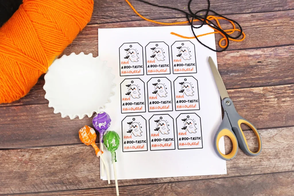 Sheet of Halloween gift tags featuring a ghost illustration and text'Have a Boo-tastic Halloween!' ready for crafting, with scissors, yarn, and candy nearby.